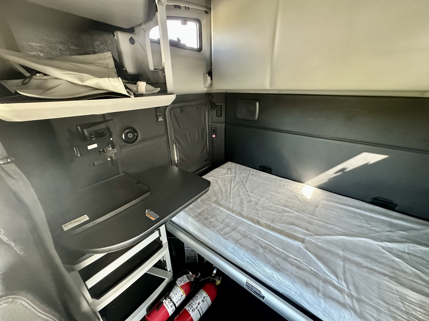 Kenworth T680 sleeper cab interior showing bunk bed with mattress, overhead storage, and control panels on left side.