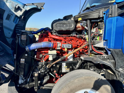 Kenworth T680 engine bay showing Cummins X15 engine with red block, blue hoses, and black air intake; components and wiring visible in cab side.