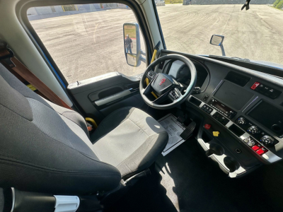 Kenworth T680 interior from driver side showing steering wheel, dashboard, pedals, and seating; side mirror visible; window view of open area.