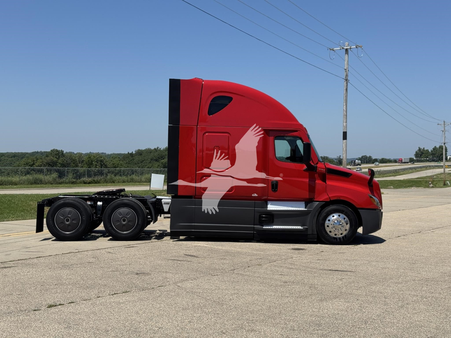 Truck side view: red 2023 Freightliner Cascadia 126 conventional sleeper with black/gray chassis and silhouette graphic on cab, 6-wheel rear, on road in open area.