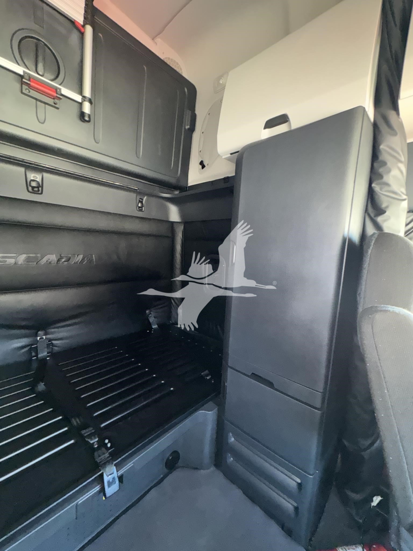 Cab interior of a Freightliner Cascadia sleeper showing storage box, overhead cabinet, and foldable bunk area with clear text panels.
