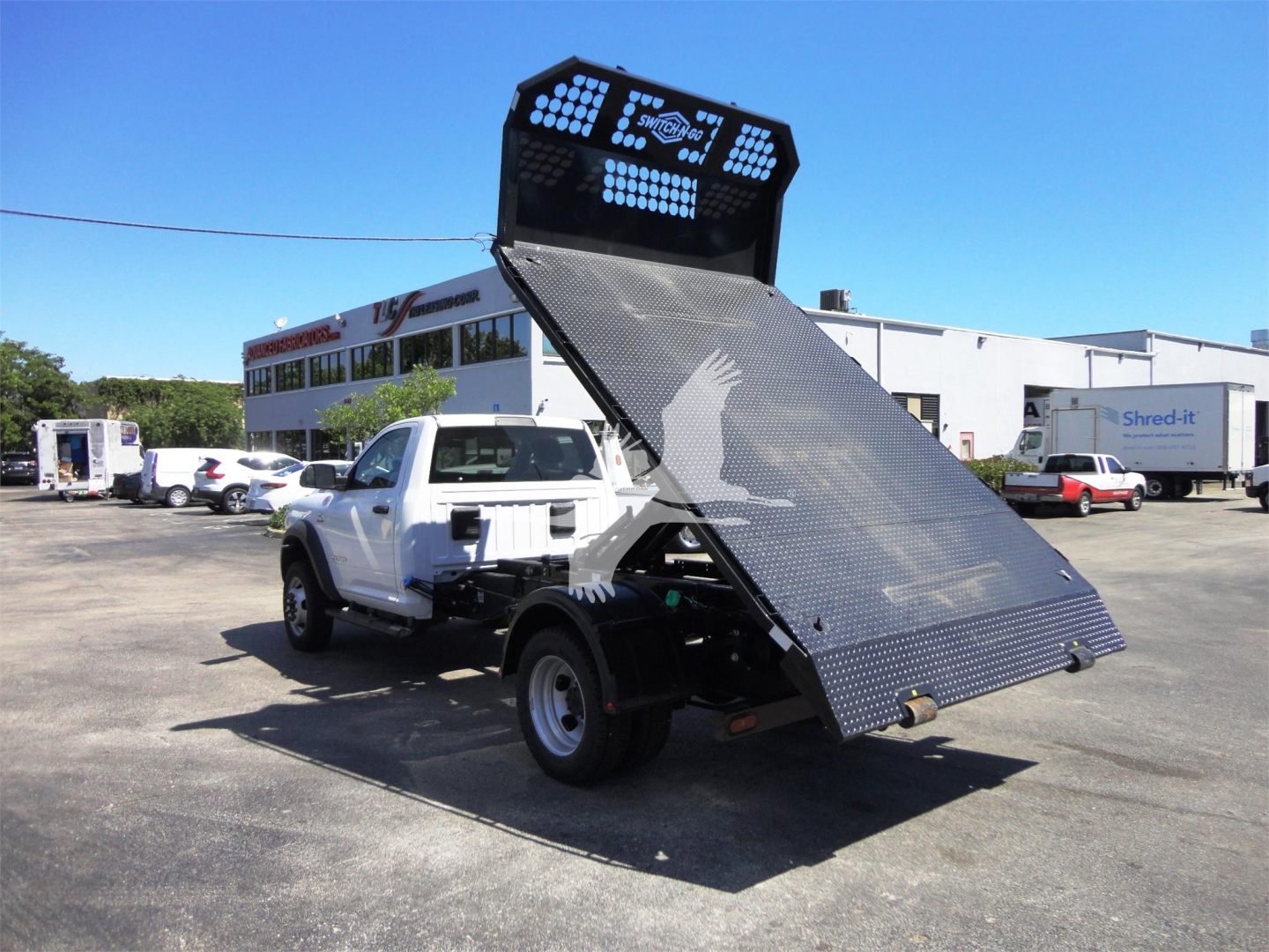 Ram 5500 dump bed or hooklift truck in a dealership lot, with a raised metal loading ramp and lighted rear LED panel.
