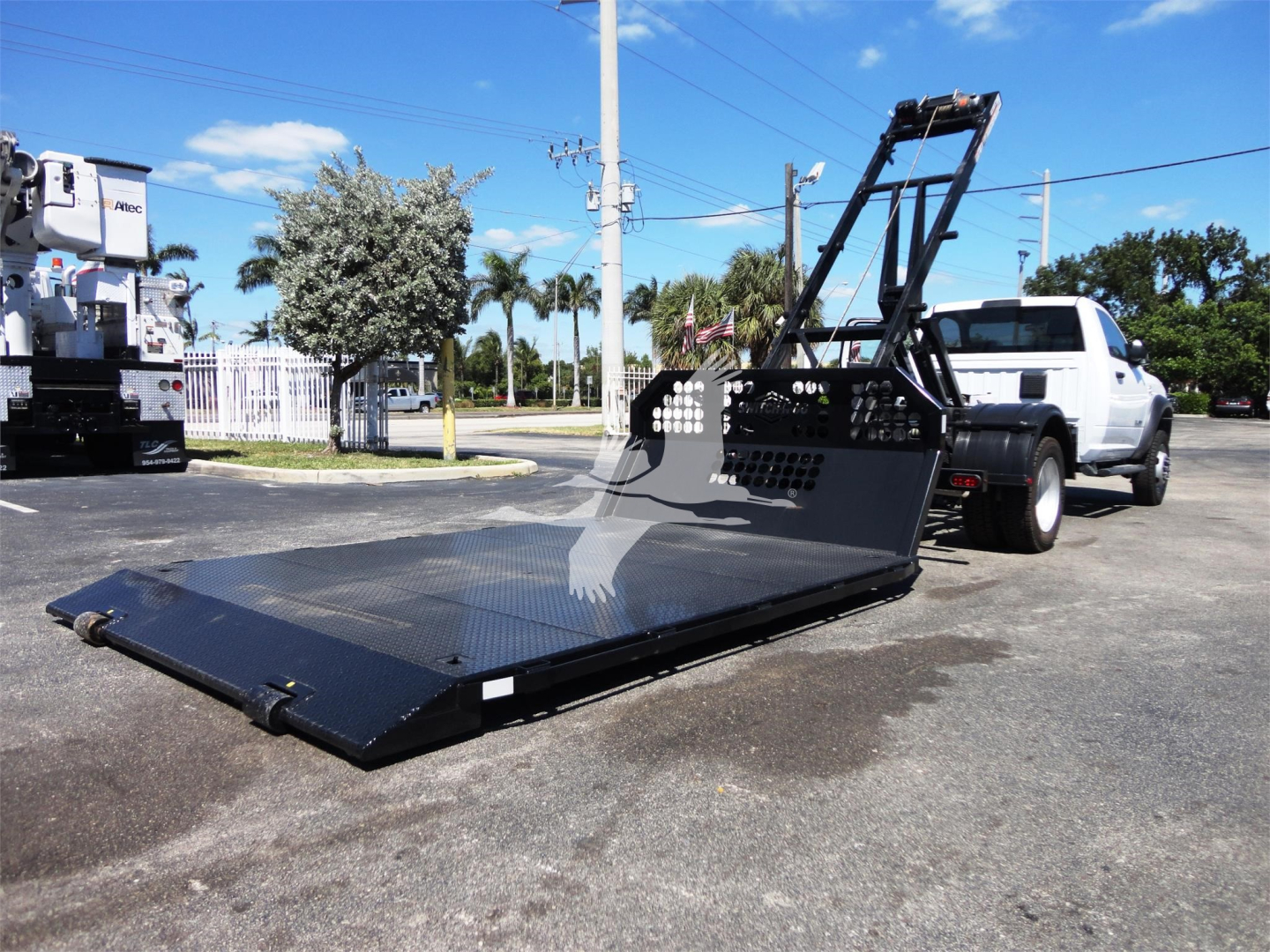 Ram 5500 Hd truck with a large black hooklift bed extended onto the ground, white cab, industrial yard background.