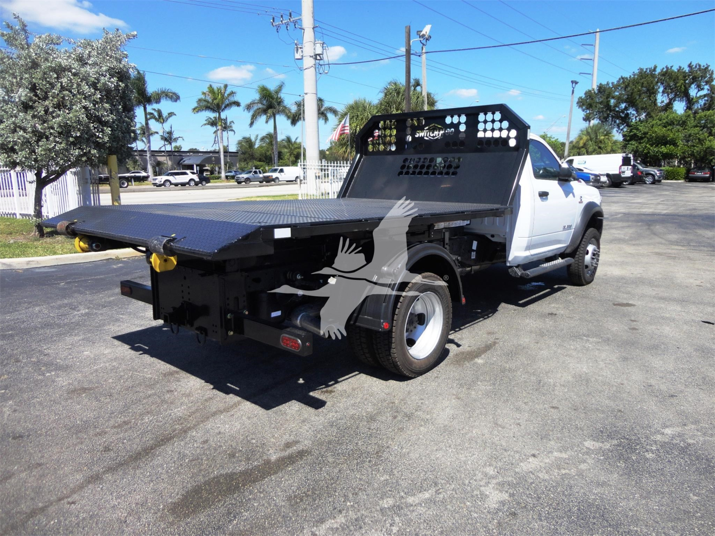 Ram 5500 Tradesman/HD hooklift truck rear view showing flatbed equipment and heavy-duty bumper, white cab, black steel ramp, and grid headache rack.