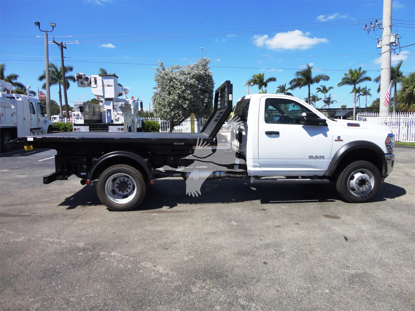 Truck side profile showing a white Ram 5500 HD with a long flatbed and hooklift equipment. Stickered details not readable from distance.