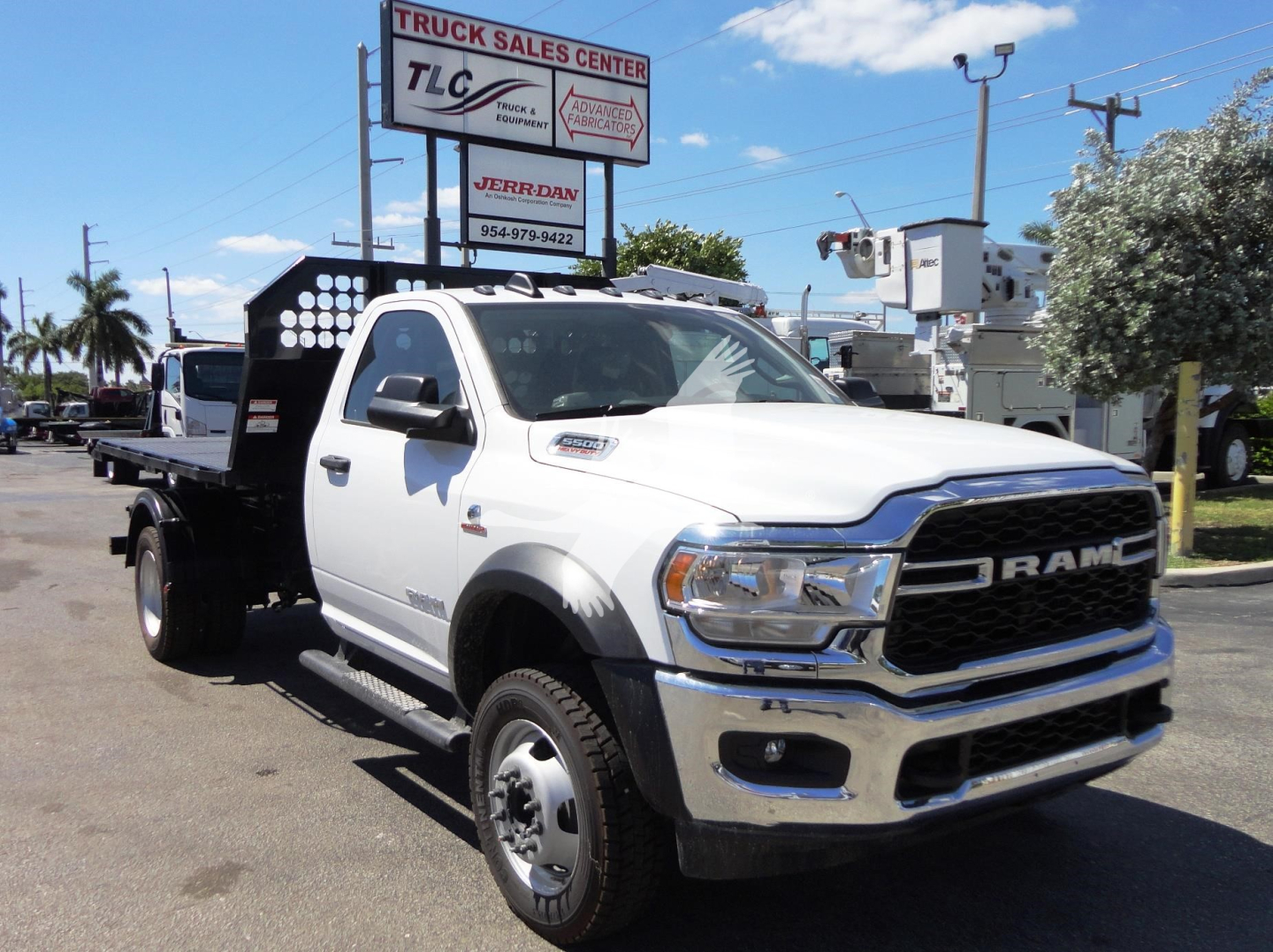 White Ram 5500 HD truck with a black hooklift bed, front grille with RAM letters, cargo area with perforated metal sides, dealership signs in background.