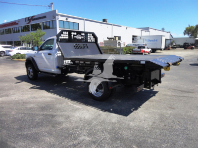 Ram 5500 HD hooklift truck showing flatbed with hydraulic mechanism and rear ramp, white cab with black equipment, dealership lot in background.