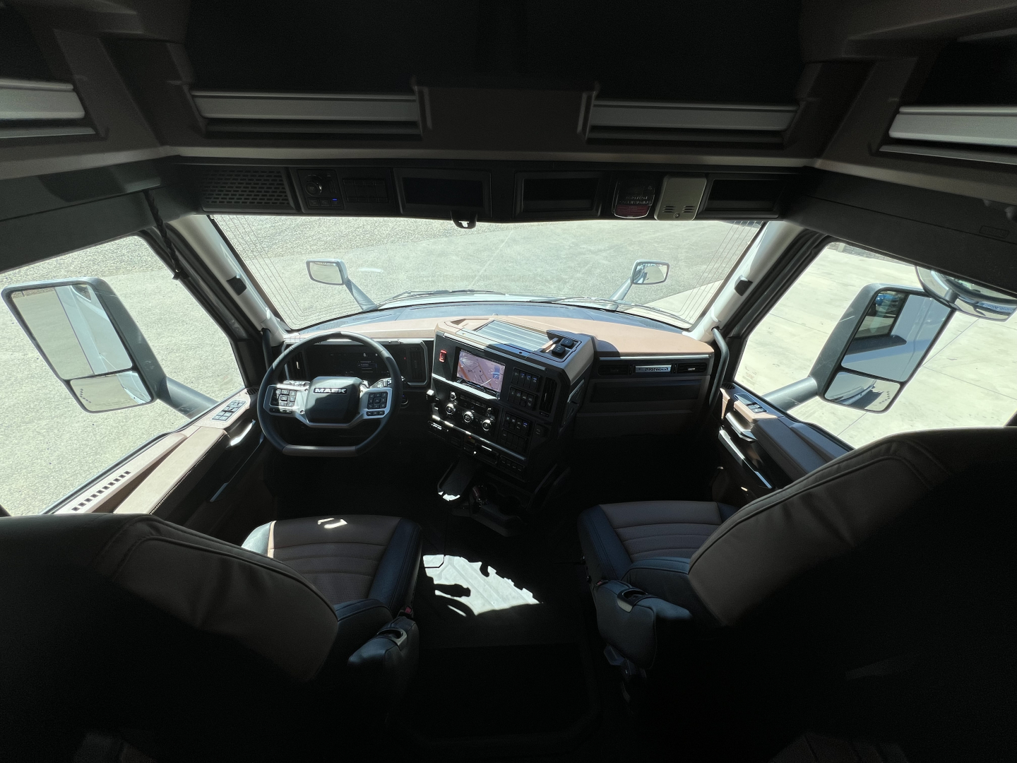 Truck cab interior view from behind windshield showing steering wheel, dashboard with central touchscreen, and two front seats in a Mack PR64T76H.