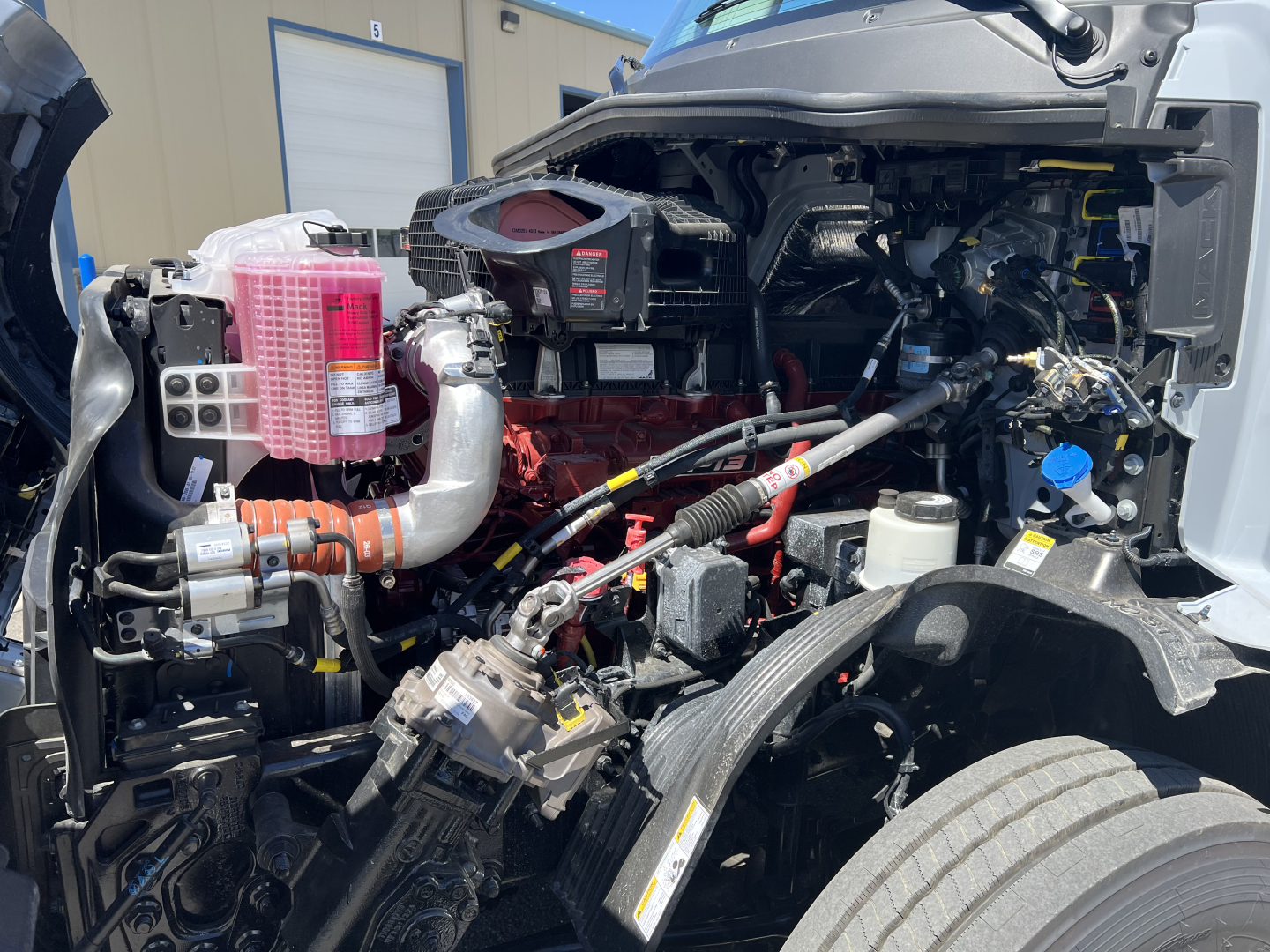 Truck engine bay of a 2027 Mack PR64T76H showing exposed engine with pink coolant reservoir, braided hoses, intake, and various components.