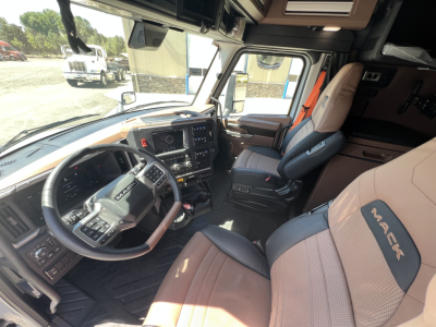 Truck cab interior of a 2027 Mack PR64T76H showing steering wheel, dashboard, driver seat with Mack logo, and passenger seat; visible window and exterior scene.