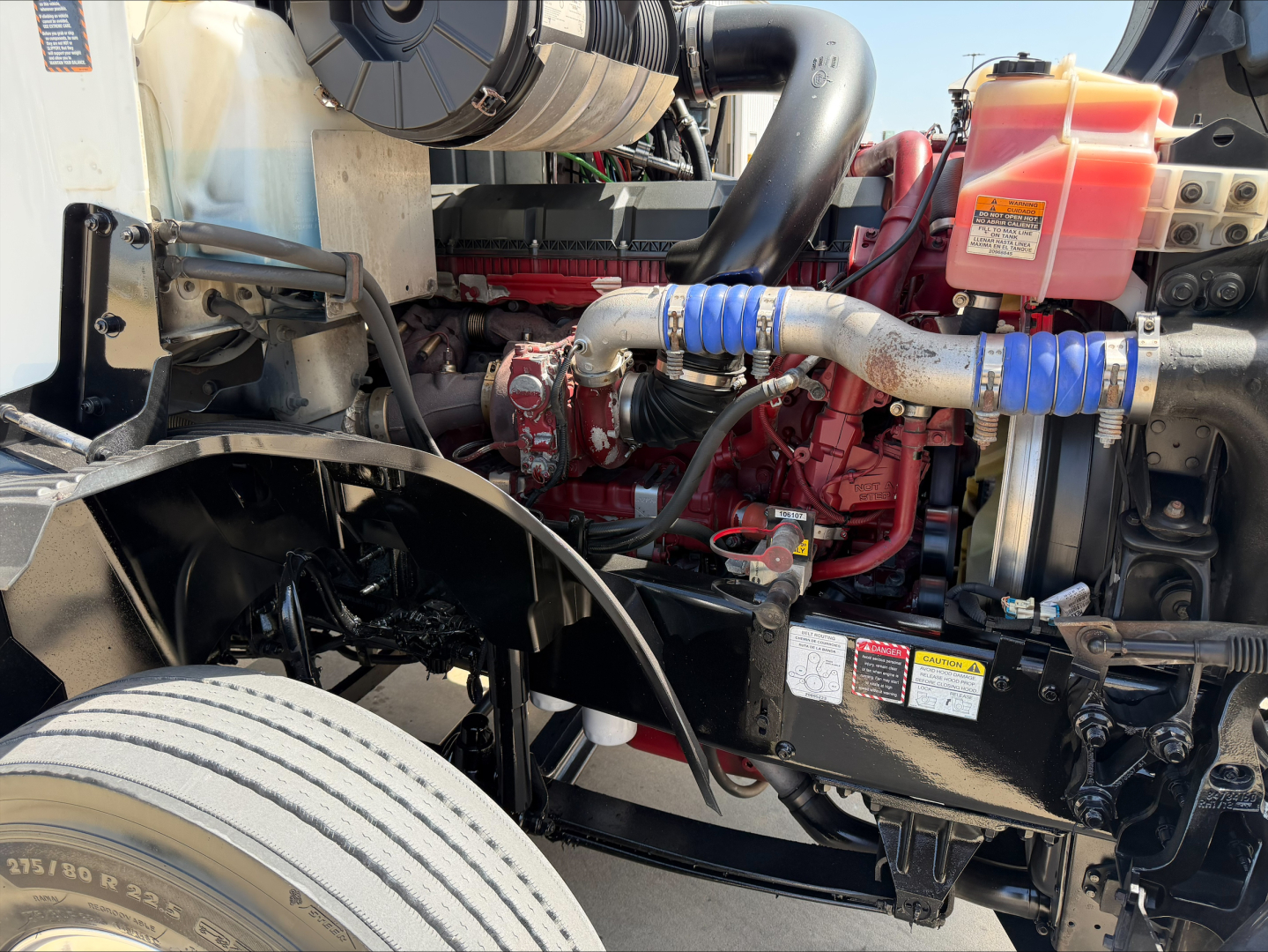 Truck engine bay view showing red diesel engine, blue hose clamps, orange coolant reservoir, and black chassis with various labels.