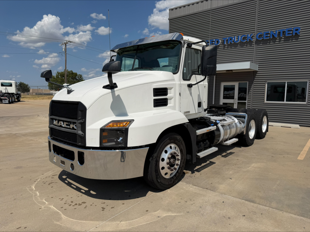 Mack Anthem conventional daycab truck in front of dealership, white cab, dual rear axles, chrome bumper, fuel tanks, grille with MACK logo.