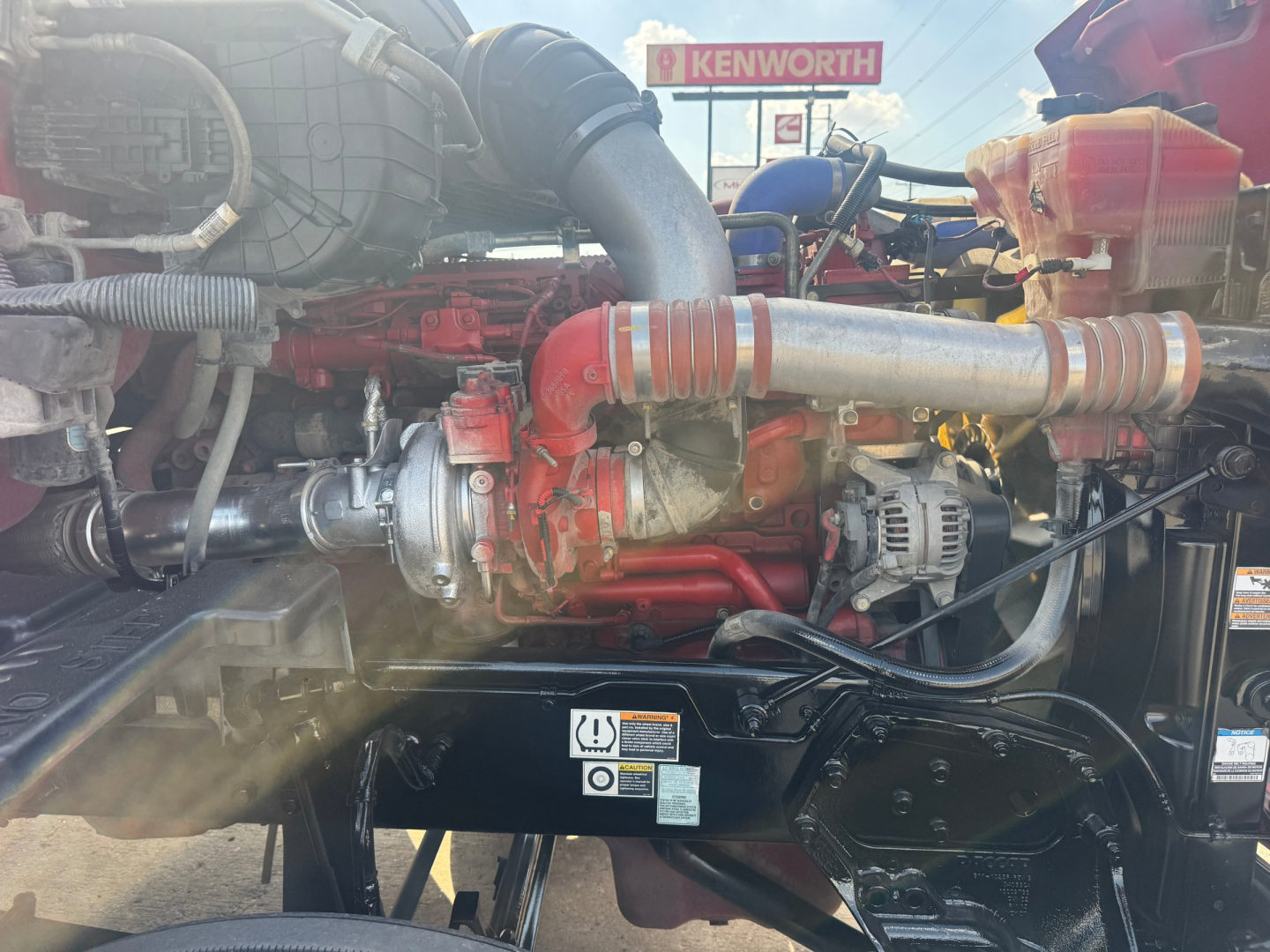 Peterbilt 579 engine bay with large red engine and turbo piping, various hoses and a Kenworth sign visible in background.