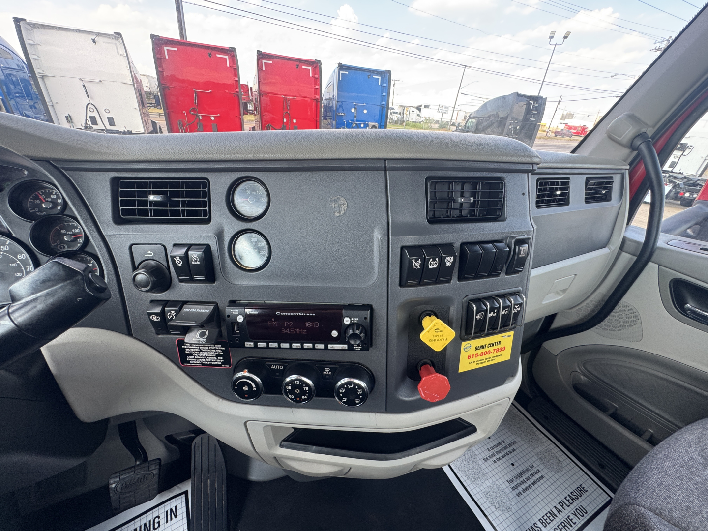Truck cab dashboard and center console in a Peterbilt 579 showing gauges, climate controls, and radio with labels; yellow tag and red knob visible.