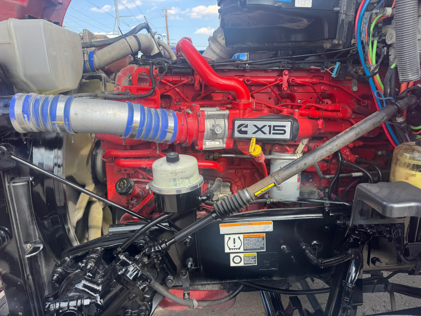 Peterbilt 579 engine bay showing red Cummins X15 engine with intake piping, coolant reservoir, and various hoses visible.