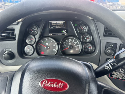 Peterbilt dashboard with multiple gauges and an infotainment screen showing a message. Steering wheel centered with Peterbilt emblem. Right stalk and various control buttons visible.