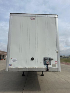 Rear view of a white Wabash box trailer showing rear doors, latches, and hitch area with support legs, small decals on lower left, and a red reflective sticker.
