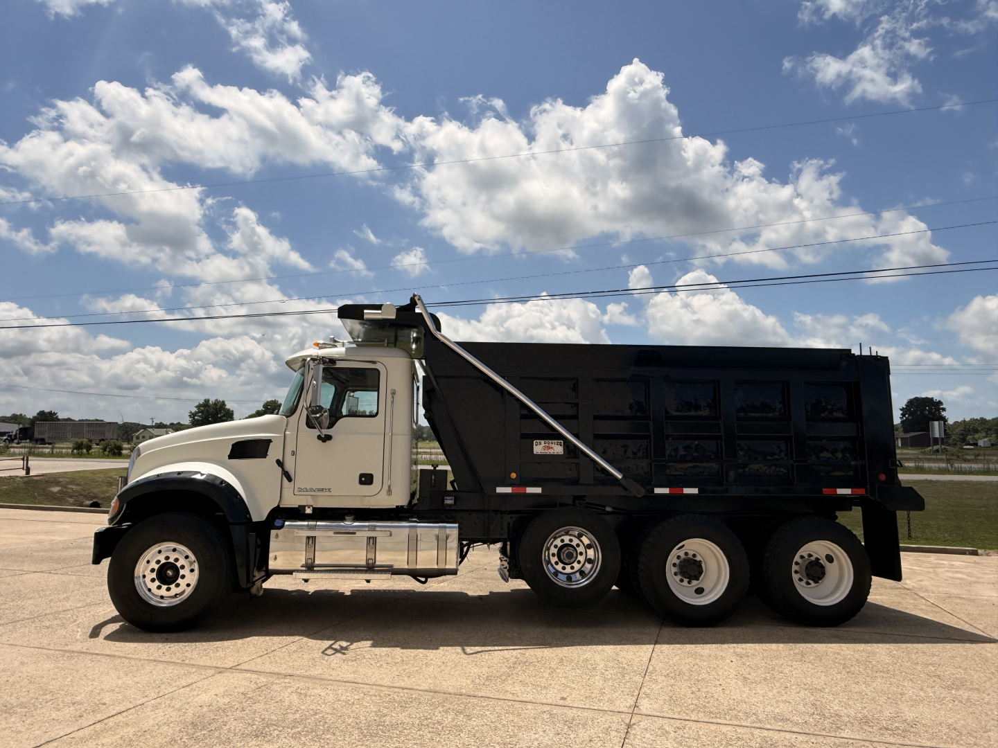 Dump truck side view showing a white cab and black dump bed with triple rear axles on a Mack GU713 chassis.