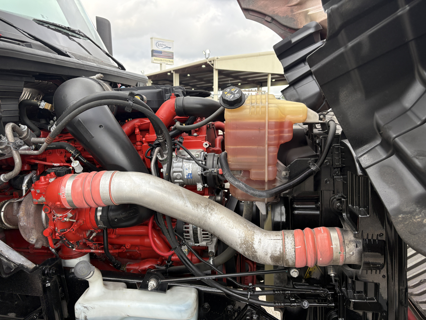 Truck engine bay showing red engine block, large silver exhaust pipe, coolant reservoir, and various hoses.