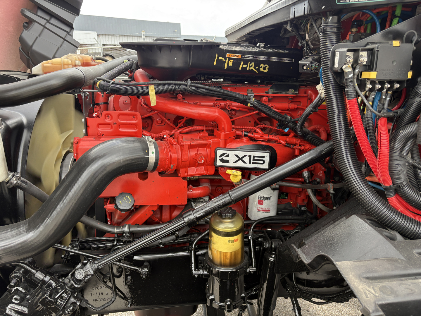 Truck engine bay showing Cummins/C-AXIS diesel engine with red valve covers, large intercooler hose, yellow oil filter, and various hoses and wiring in front of a sleeper truck.