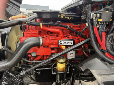 Truck engine bay showing Cummins/C-AXIS diesel engine with red valve covers, large intercooler hose, yellow oil filter, and various hoses and wiring in front of a sleeper truck.