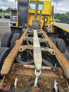 Truck chassis and cab frame showing rusty frame rails and fifth wheel mounting with yellow cab in background.