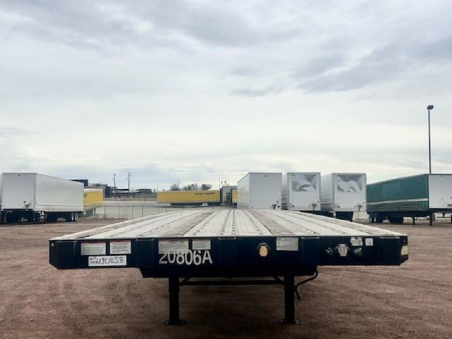 Flatbed trailer in a lot with other trailers in the background; front view showing black metal deck with white stenciled numbers 20806A and a license plate.