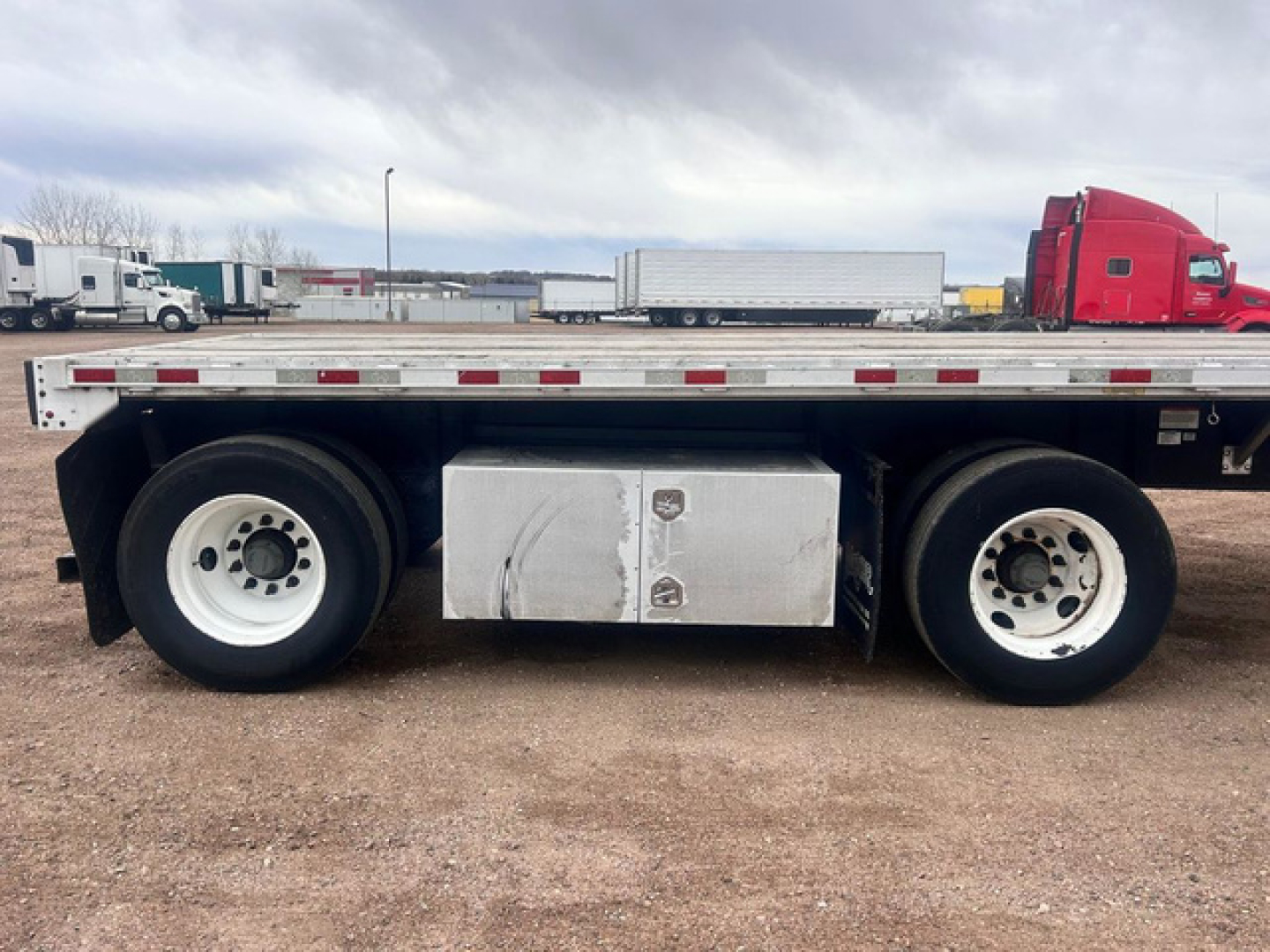 Flatbed trailer side view showing dual rear axles with white wheels and metal toolbox mounted between axles.