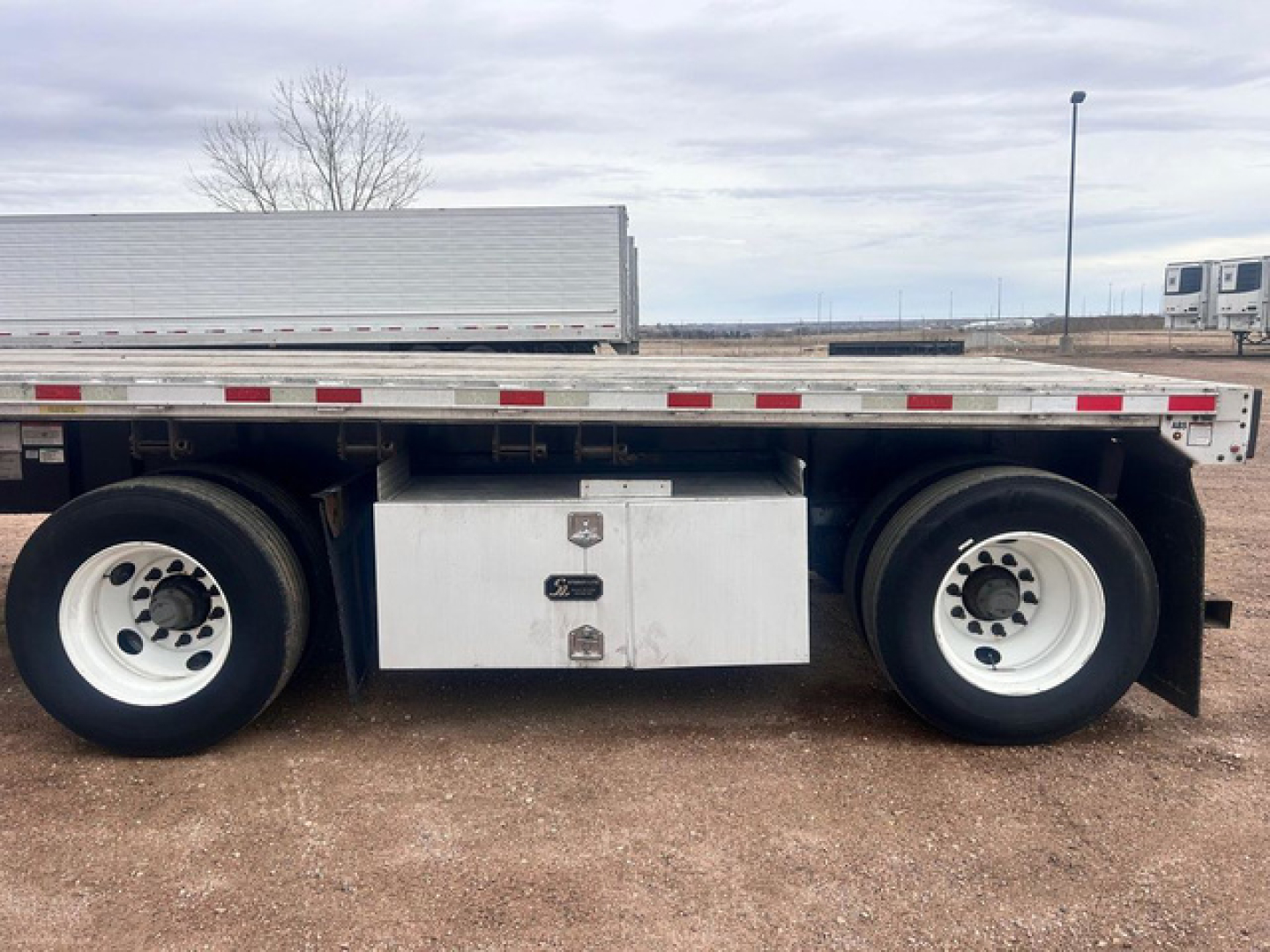 Flatbed trailer side view showing dual rear wheel axles and aluminum tool/storage box beneath the bed.