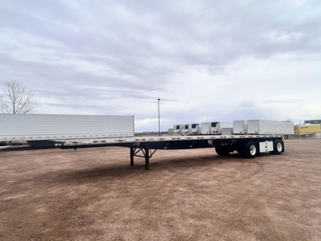 Flatbed trailer in a dirt lot showing a long, low profile with a swing-out gooseneck and multiple axles at the rear.