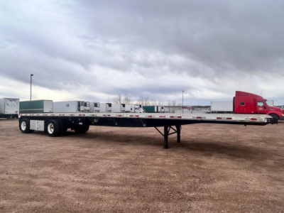 Flatbed trailer in a lot with red truck cab in background; long lowboy with support legs down, various equipment boxes near front.