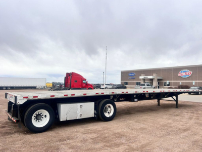 Flatbed trailer showing dual rear axles and white storage box, long metal bed with stake pockets, overcast sky, dealership lot in background.