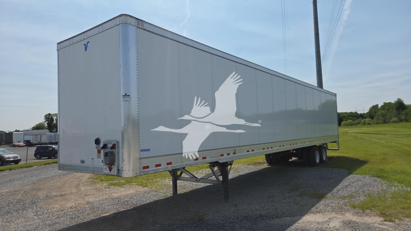 Vanguard VXP trailer side view showing a gray cargo trailer with a white crane graphic, undercarriage legs and wheelset, no cab in sight.