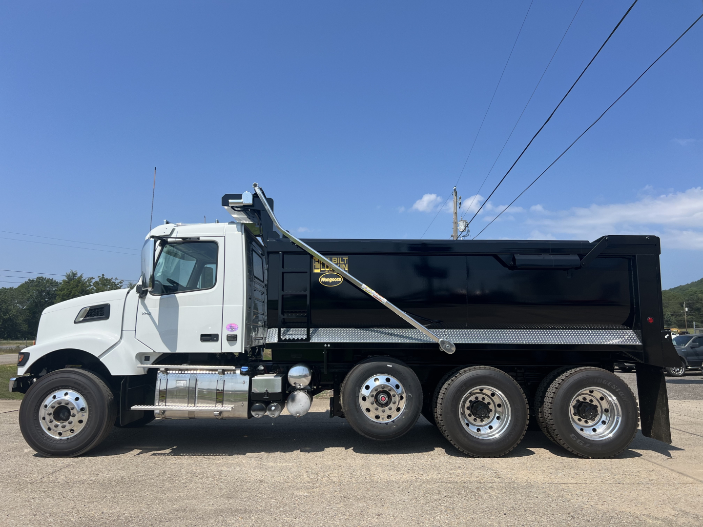Dump truck: side profile of a white Volvo VHD64F300 dump truck with black dump body. Visible text: no text clearly readable on truck besides generic labels; overall vehicle details: large wheelbases, dual rear axles, chrome fuel tanks and s