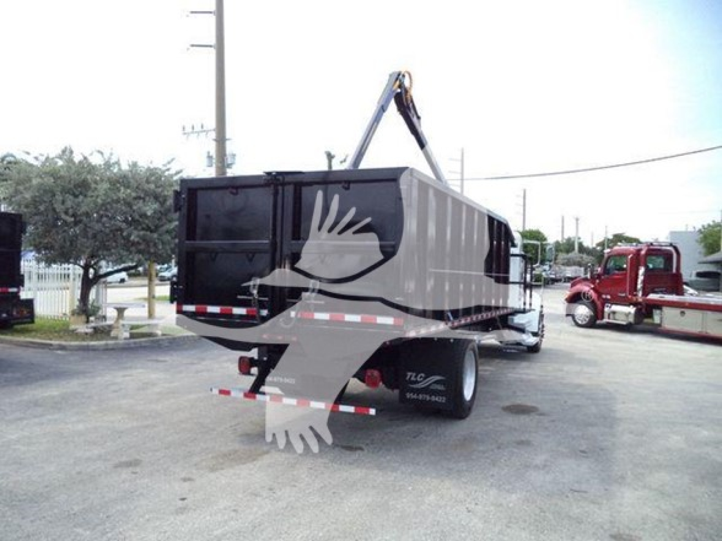 Truck body with grapple hooks lifting a black dump/dump-style trailer in a dealership yard. Visible markings: no legible labels on the side; rear shows reflective stripes.