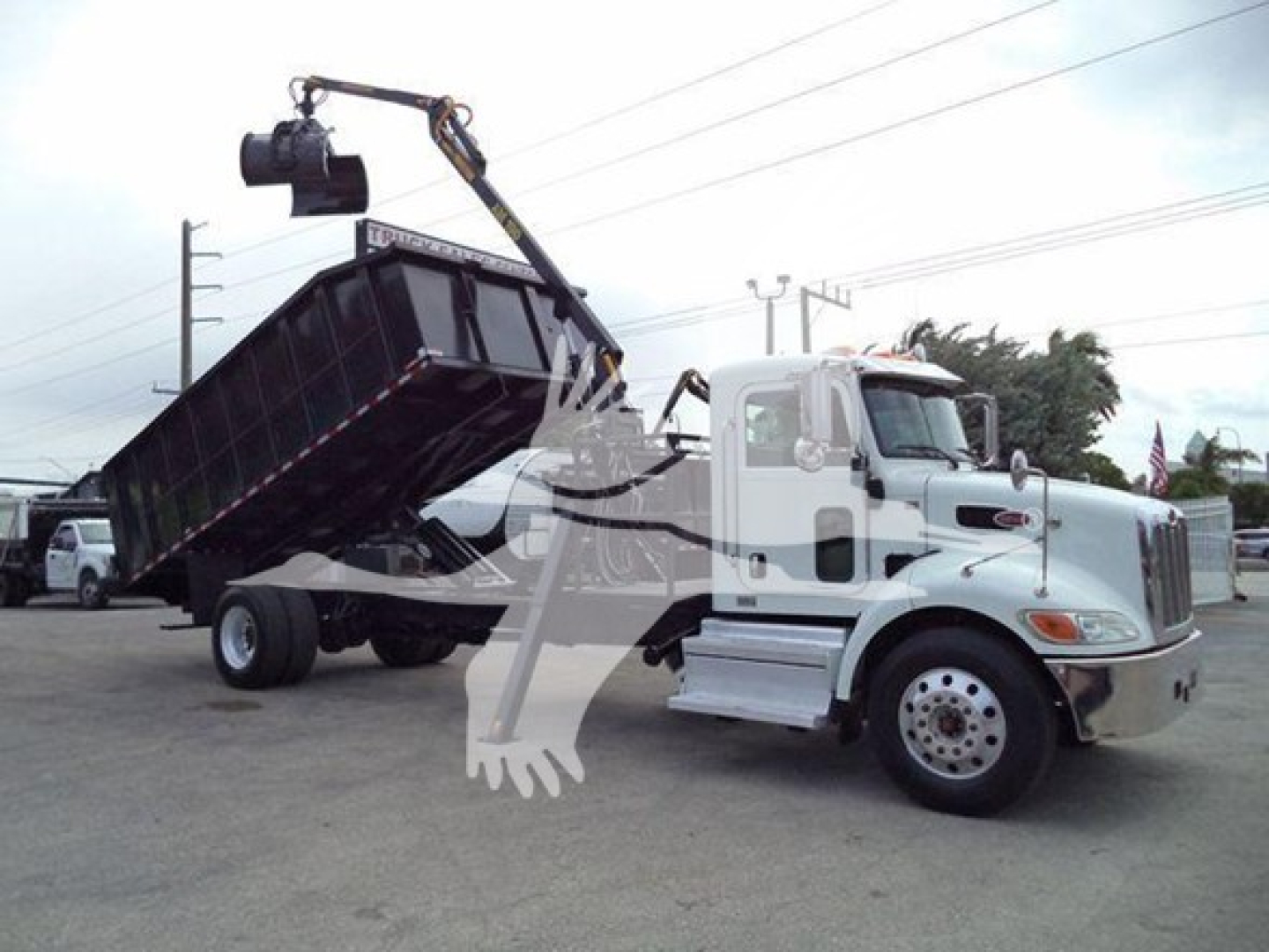 Peterbilt 337 truck with a tilted grapple bed in a yard, white cab, black dump body, crane arm overhead lifting a bucket