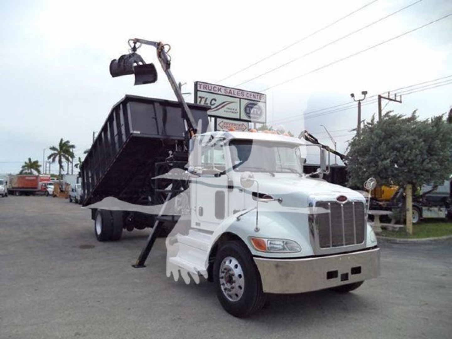 Peterbilt 337 truck with a lifted grapple/dump bed in a dealership lot. Visible text on sign in background: 'TRUCK SALES CENTER TLC' and on truck cab area there are no legible model details.
