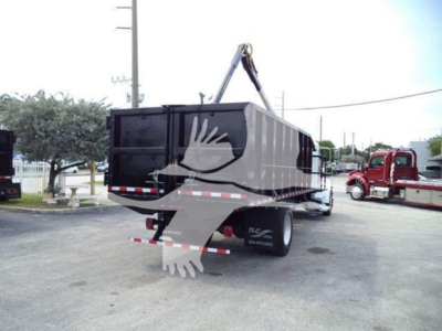 Truck body with grapple hooks lifting a black dump/dump-style trailer in a dealership yard. Visible markings: no legible labels on the side; rear shows reflective stripes.