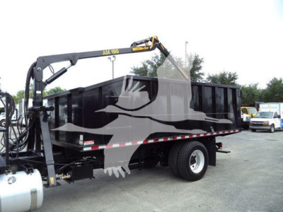 Truck bed with hydraulic grappler arm; side shows black dump bed on Peterbilt chassis; visible label on crane reads AV 90 (partial) and hydraulic hoses at front.