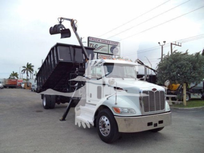 Peterbilt 337 truck with a lifted grapple/dump bed in a dealership lot. Visible text on sign in background: 'TRUCK SALES CENTER TLC' and on truck cab area there are no legible model details.