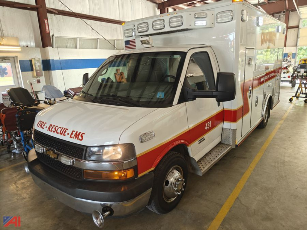 Ambulance front-left view showing 2015 Chevrolet Express G4500 EMS ambulance with white body, orange-red stripe, emergency lights on cab and roof, and 'FIRE-RESCUE-EMS' text on the front bumper.