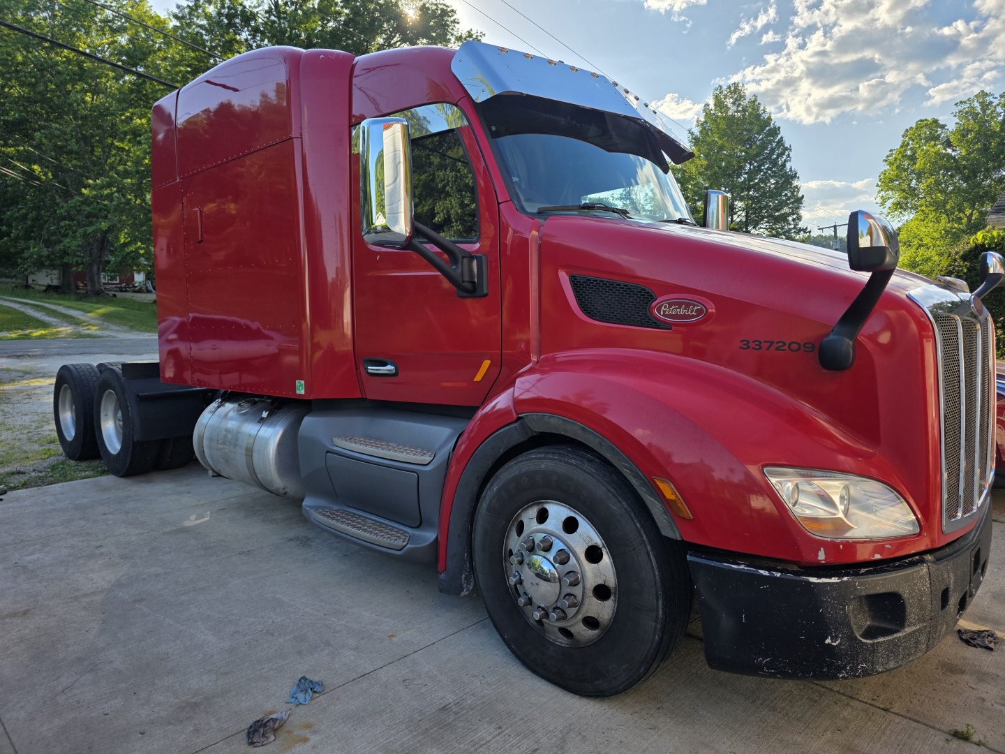 Peterbilt 579 semi-truck cabin in red, side view, with dual rear axles and chrome fuel tanks, green trees in background.
