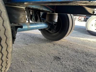 Underbelly view of a trailer axle and wheel assembly, blue frame, visible suspension components and tire tread.