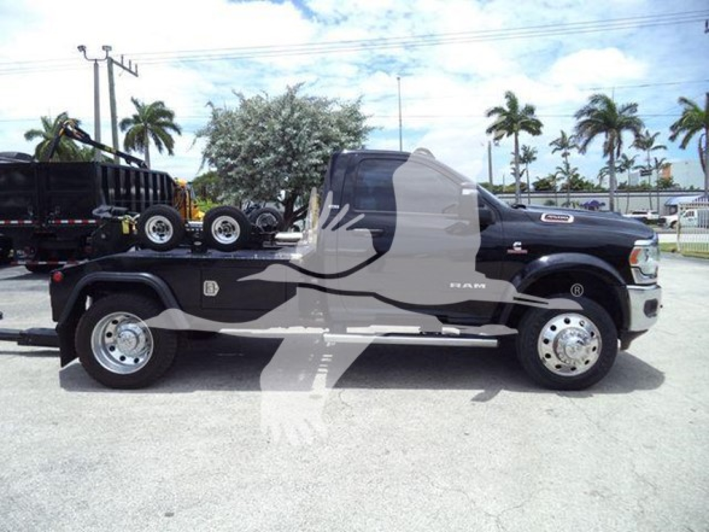 Side view of a black 2024 Ram 4500 pickup with a long bed and winch equipment; visible badges: RAM on door, chrome wheels; flatbed with dual wheels on trailer.