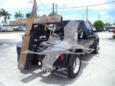 Truck bed with heavy equipment and a winch attachment mounted; side of trailer shows phone numbers and towing decals; wheel and fender visible.