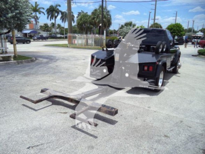 Ram 4500 truck with winch setup; two long curved hitch-like bars and a metal winch sled on ground; black cab with spare tire mounted in bed; visible road and palm trees in background.