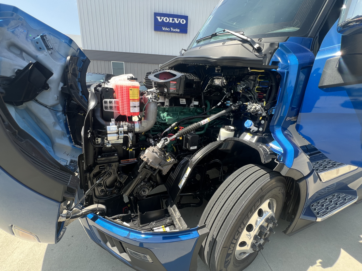 Volvo VNL64T860 engine bay with open hood showing coolant reservoir, air intake, and engine components; blue truck cab in view.