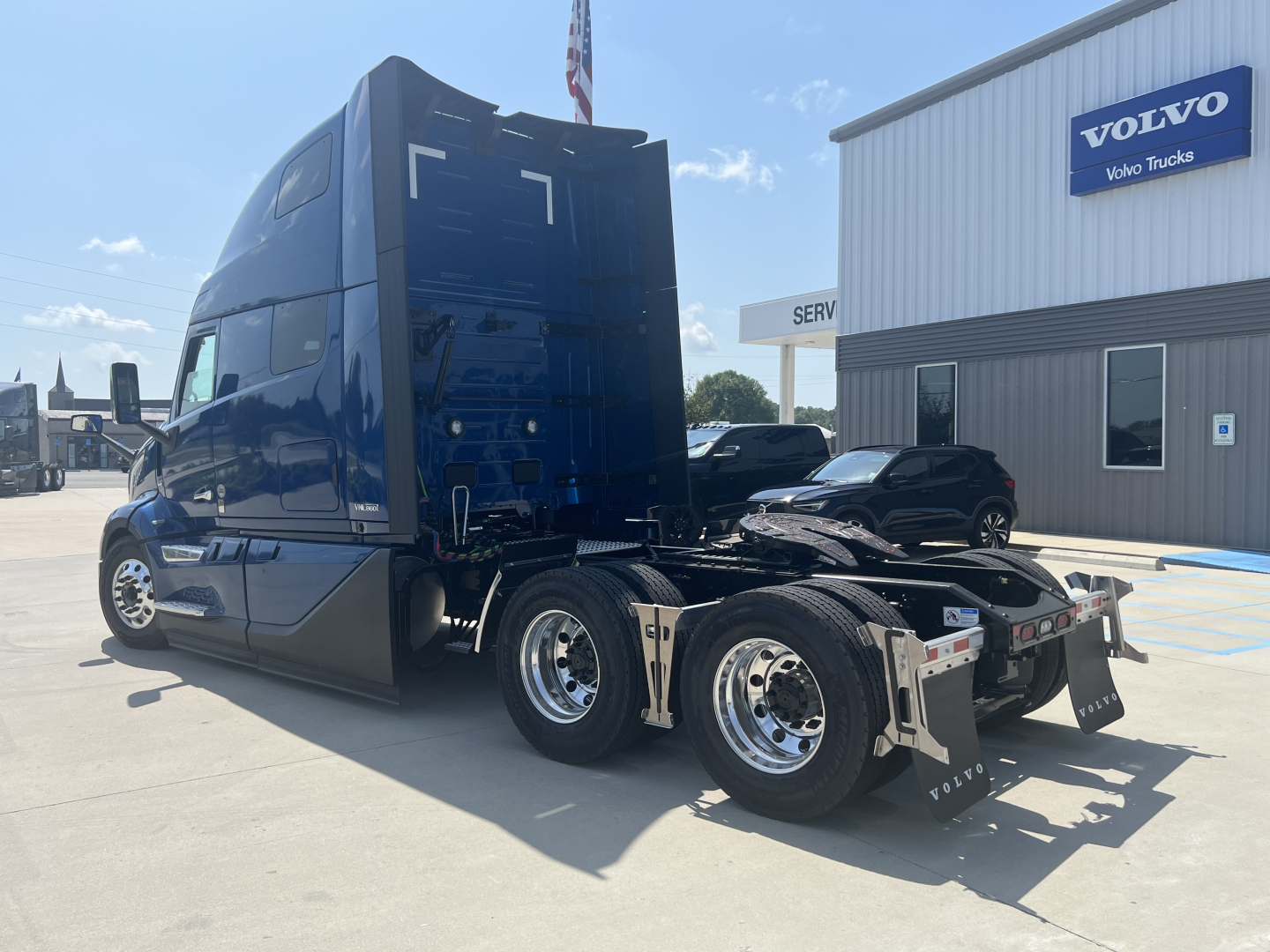 Side view of a blue Volvo VNL truck tractor with sleeper cab, showcasing rear wheels and fifth wheel area, dealership building in background with Volvo sign.