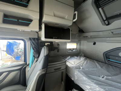 Interior of a Volvo VNL64T860 sleeper cab showing overhead storage compartments, a small monitor, window, and sleeping area with plastic-wrapped mattress.