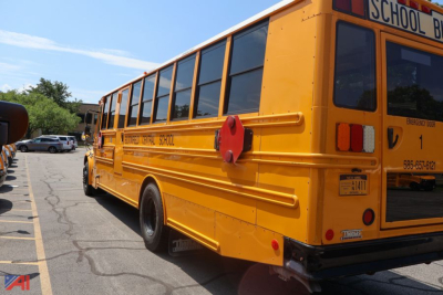 Rear and side view of a yellow school bus with black-tinted windows. Visible text includes 'SCHOOL BUS' on the rear window, a red circular safety device on the side, and small license/inspection stickers near the back; the bus has black bum